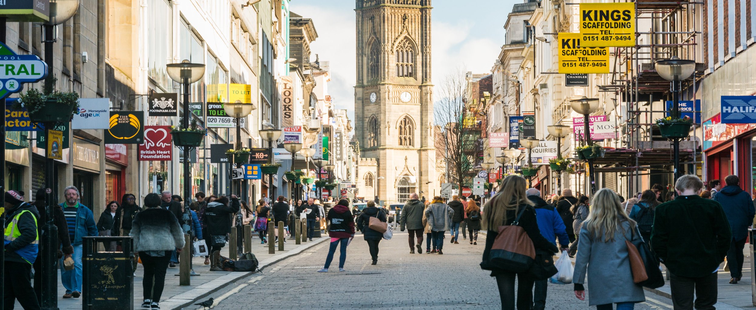 Bustling city street with pedestrians, shops, and a prominent clock tower in the distance.