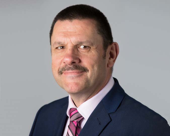 Headshot of a man with a mustache, wearing a blue suit and pink striped tie, against a gray background.