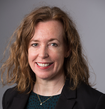 Professional headshot of a smiling woman with wavy brown hair, wearing a dark blazer and a green patterned top.