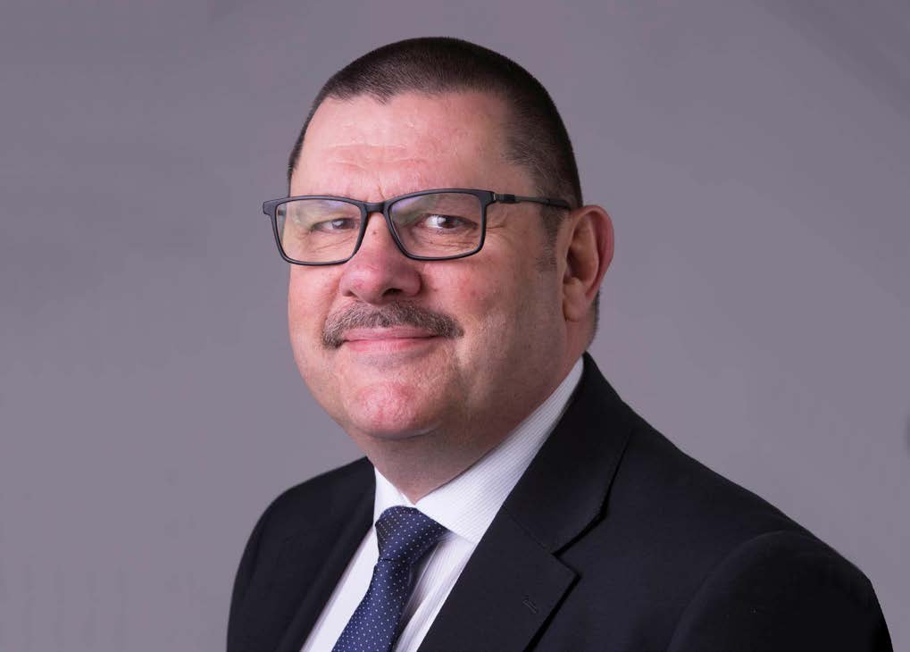 Headshot of a man in a dark suit, white shirt, and patterned tie, wearing glasses and a mustache, smiling slightly.