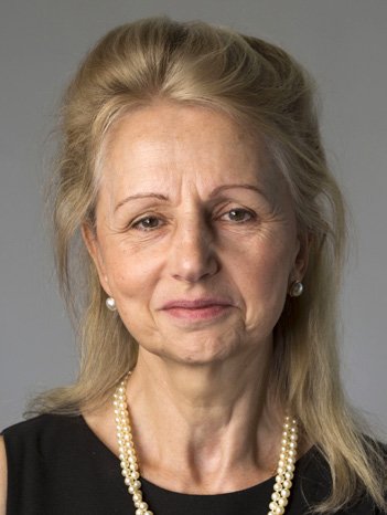 Portrait of a blonde woman wearing a black top and multiple strands of pearl necklaces, looking directly at the viewer.