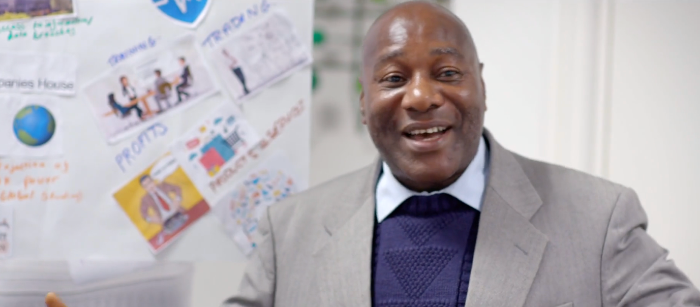 Man in a grey suit and blue sweater smiling, with a whiteboard behind him featuring various small diagrams and text related to business topics.