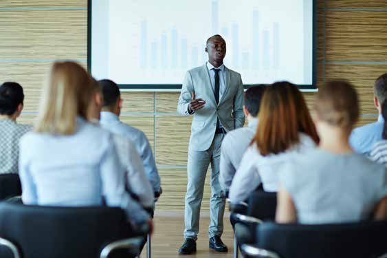 A man in a suit presenting to an audience, with a bar chart displayed on a projector screen behind him.