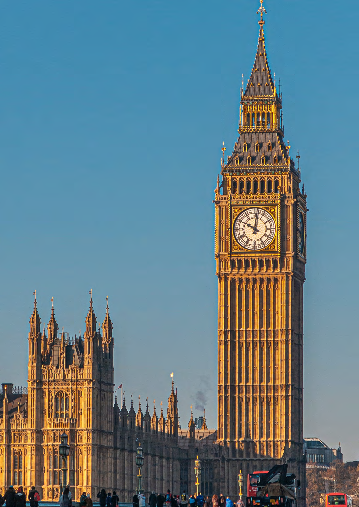 Big Ben clock tower and the Houses of Parliament in London under a clear blue sky, with people and red buses visible on a bridge in the foreground.