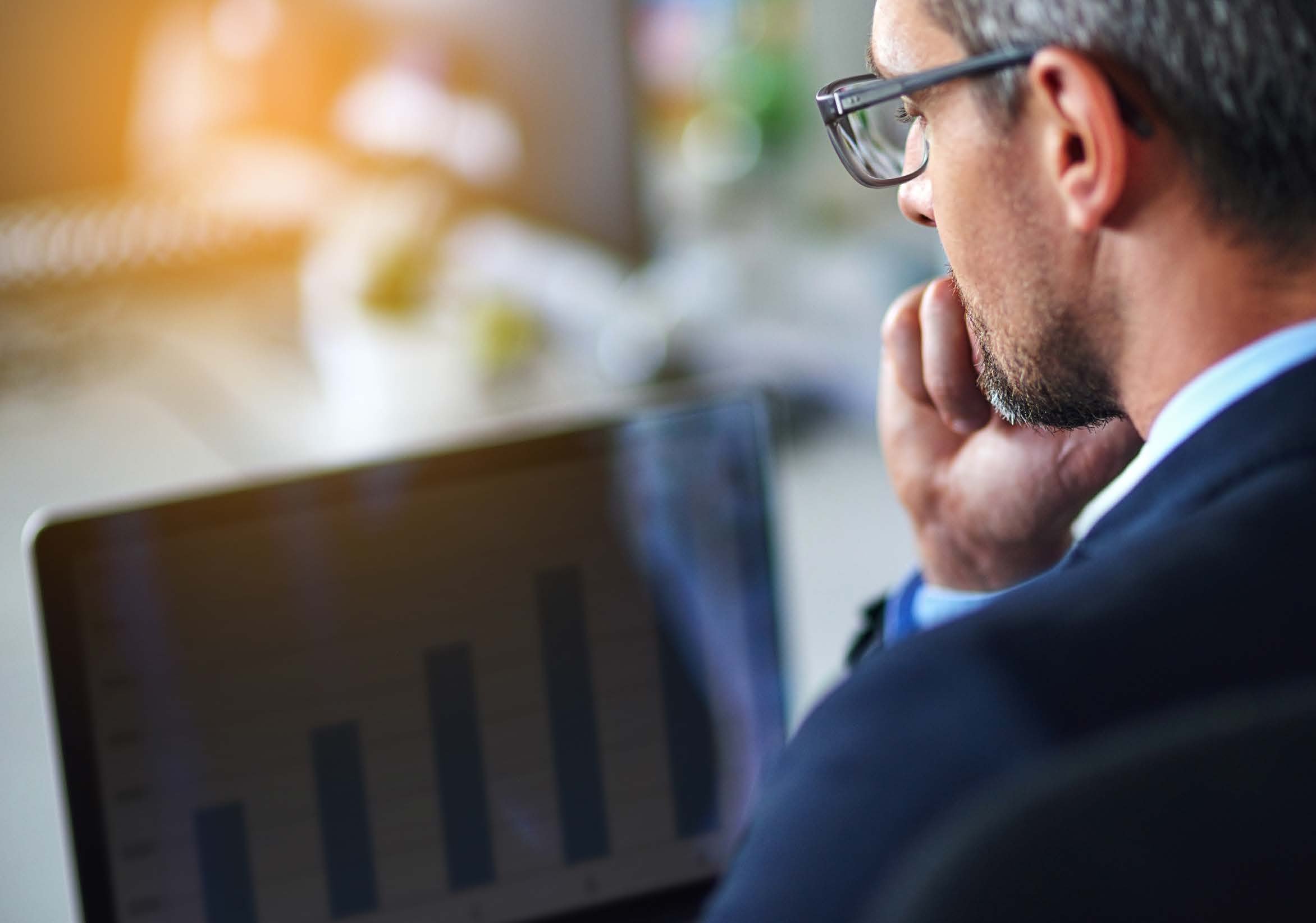 A man in glasses looking intently at a laptop screen displaying a bar chart, suggesting data analysis.