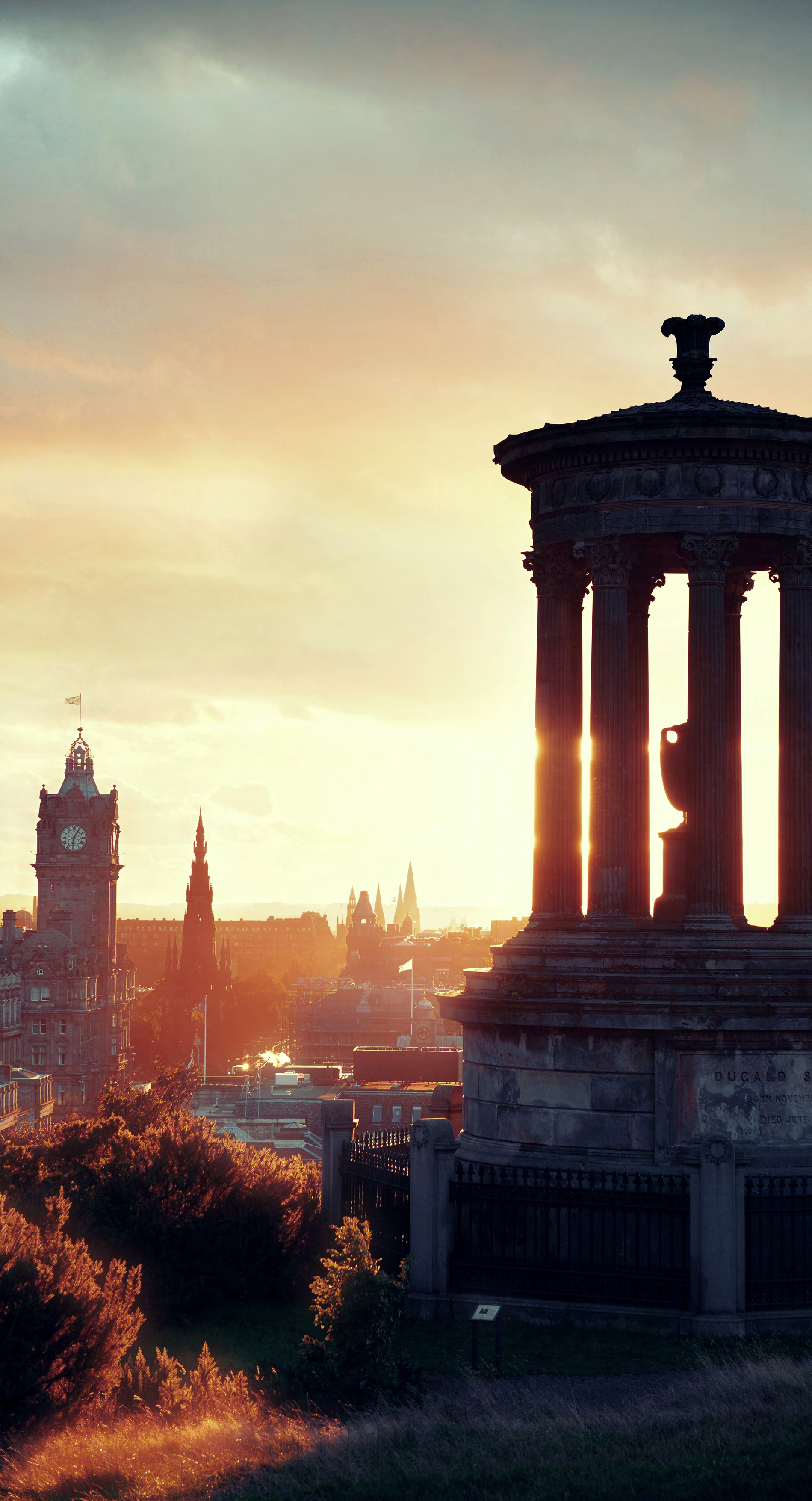 Sunset view of Edinburgh cityscape featuring the Dugald Stewart Monument and Balmoral Hotel clock tower.