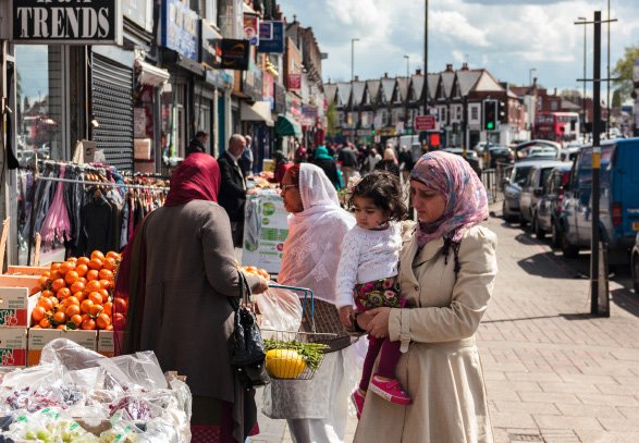 A street scene with people walking, market stalls selling fresh produce, and shops lining the street in a busy urban environment.