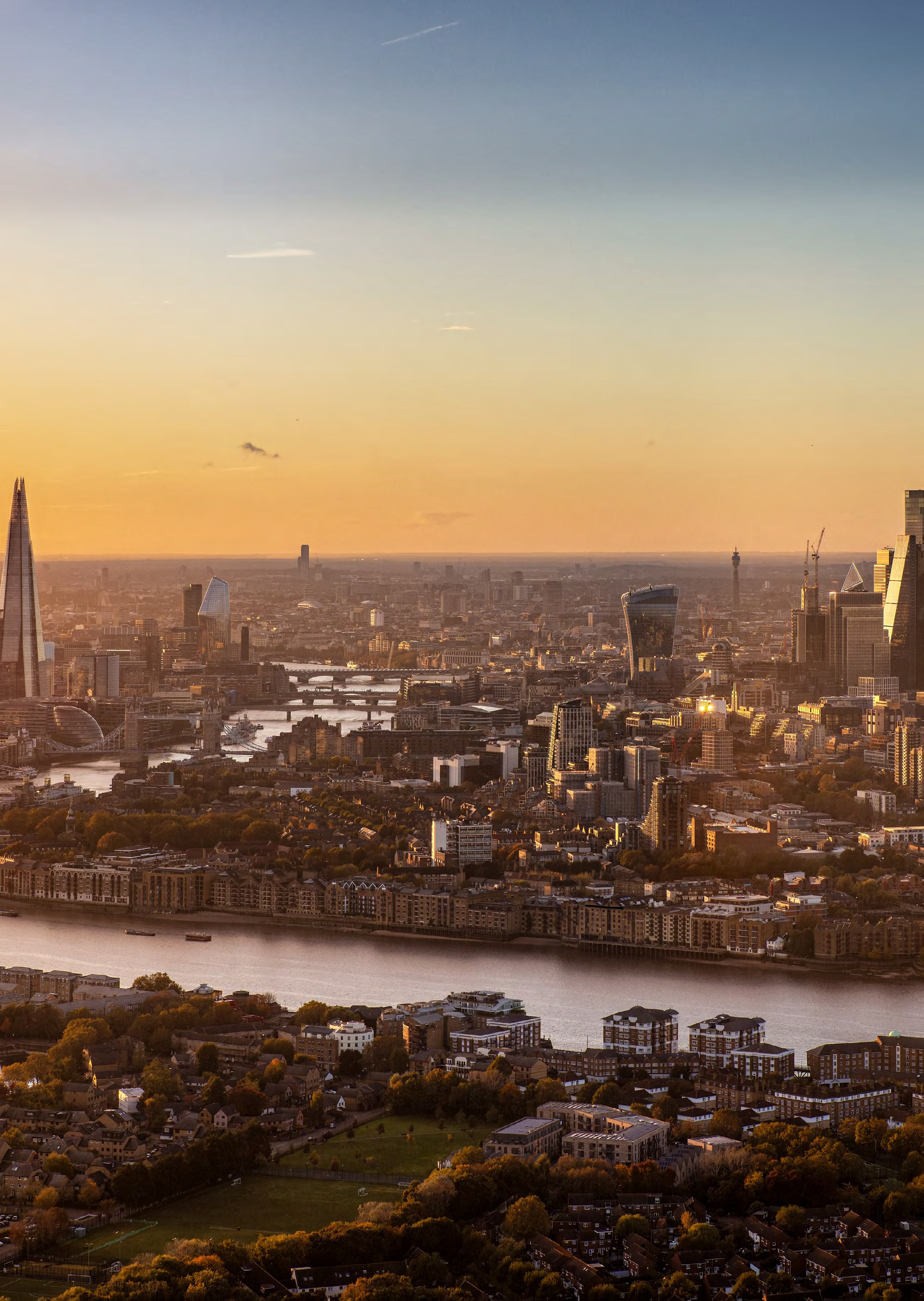 Aerial view of a vast city skyline at sunset with a river, numerous buildings including The Shard, and a warm glow.