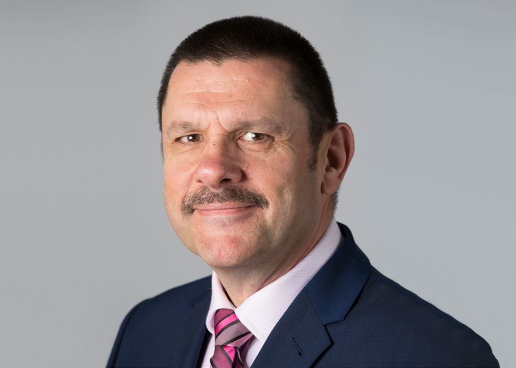 Headshot of a man with a mustache wearing a dark suit and a pink striped tie, smiling slightly against a plain gray background.