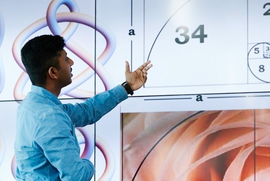 A man points to a large screen displaying mathematical concepts, including a Fibonacci spiral, numbers, and geometric patterns.