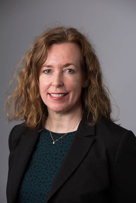 Headshot of a smiling woman with wavy auburn hair, wearing a black blazer and patterned green top.