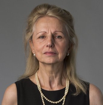 Headshot of a mature woman with blonde hair, wearing a black dress and pearl necklaces.