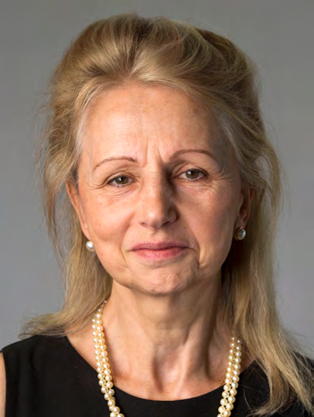 Headshot of an older woman with blonde hair, wearing a black top and pearl necklaces and earrings, looking directly at the camera.