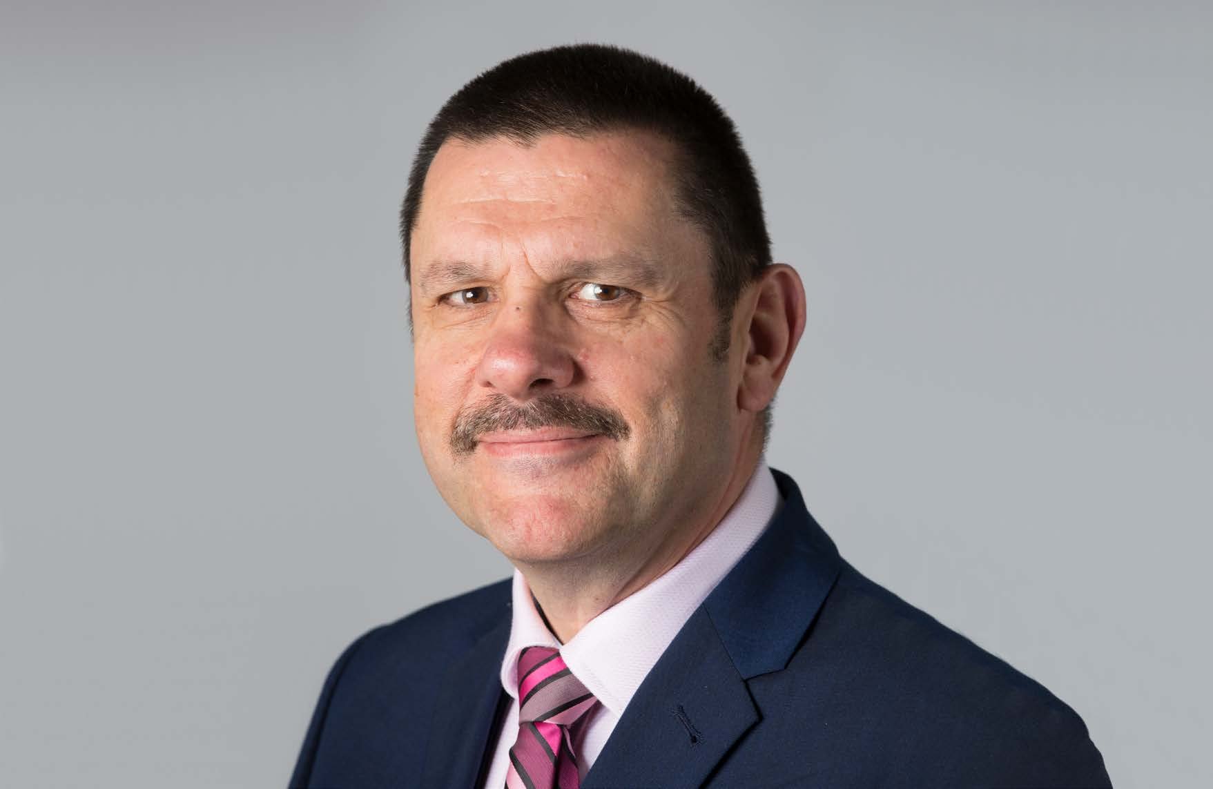 Headshot of a man with a mustache, wearing a dark blue suit and a pink striped tie.