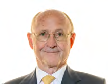Headshot of an older man with glasses, wearing a suit and yellow tie, smiling slightly, against a white background.
