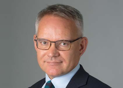 Headshot of a man with short grey hair and glasses, wearing a dark suit and a striped tie, smiling slightly.