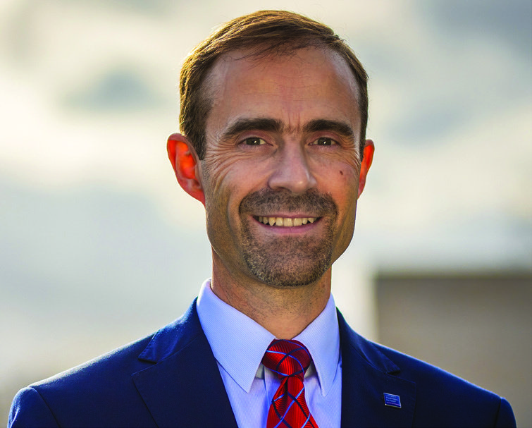 Headshot of a smiling man with short dark hair and a short beard, wearing a blue suit and red patterned tie.