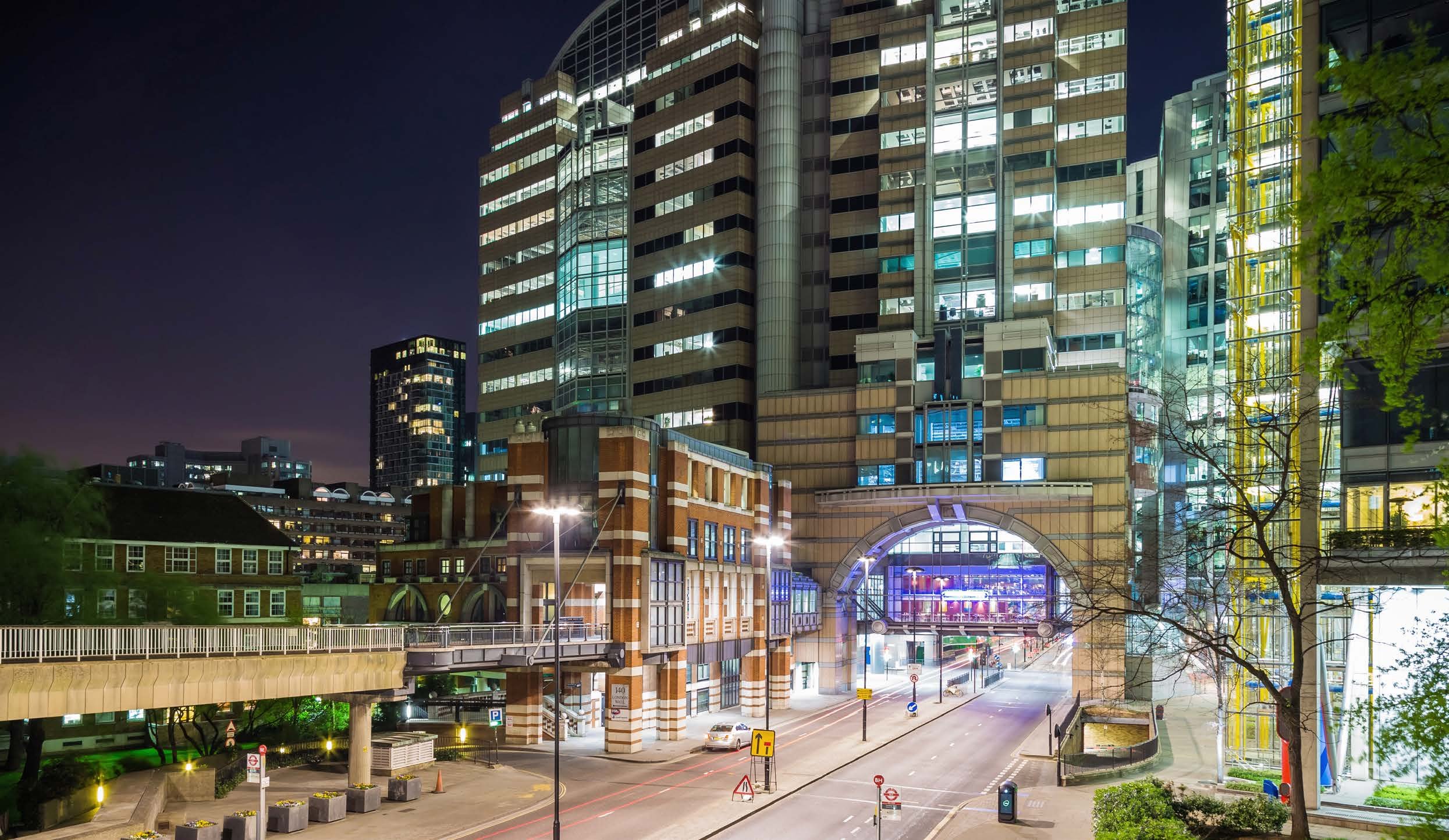 Night view of modern city buildings with illuminated windows, a street with car trails, and an elevated pedestrian bridge.