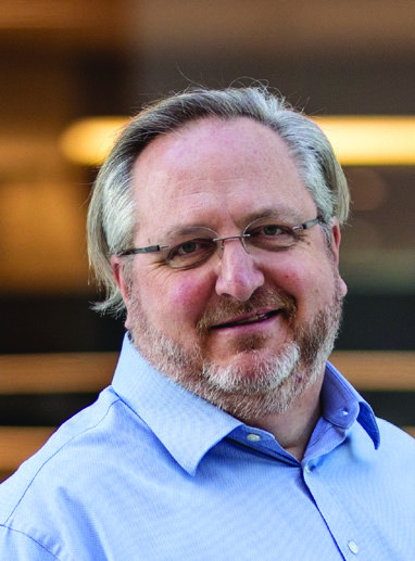 Headshot of a smiling man with glasses, graying hair and beard, wearing a light blue collared shirt.