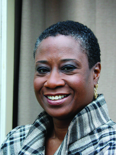 Headshot of a smiling woman with short dark curly hair and earrings, wearing a black and white plaid coat.