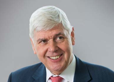 Headshot of an older man with white hair smiling, wearing a blue suit and red tie.