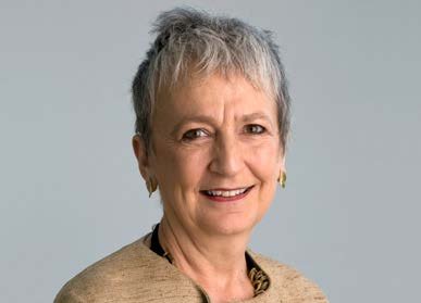 Headshot of an older woman with short grey hair smiling, wearing a tan jacket and gold earrings.