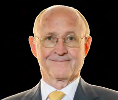 Headshot of an older man with glasses, wearing a suit and yellow tie, smiling slightly.