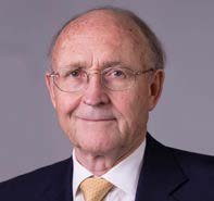 Headshot of an older gentleman with glasses, wearing a dark suit and yellow patterned tie, smiling.